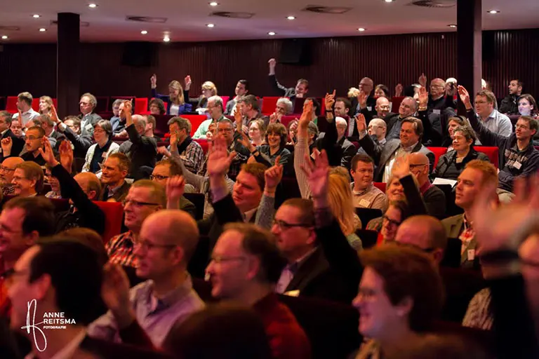 Een volle zaal waarin deelnemers hun hand opsteken