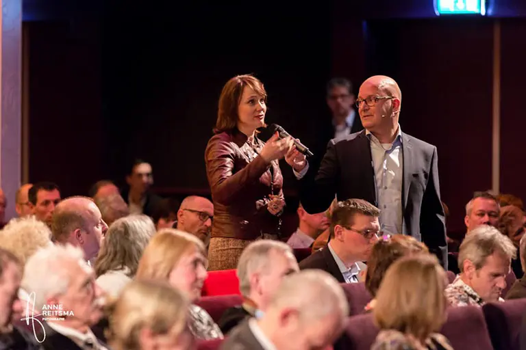 Een deelneemster spreekt in de microfoon tussen het publiek, met Gert-Jan Jansen naast haar