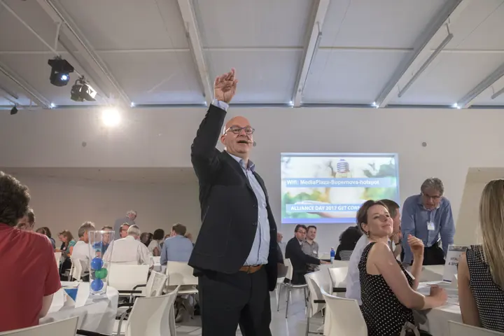 Gert-Jan Jansen steekt zijn arm op in een zaal vol tafels tijdens de Alliance Day
