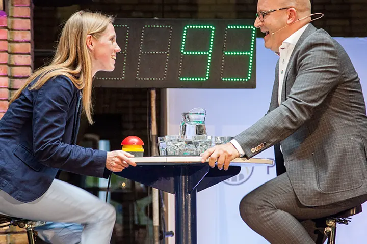 Gert-Jan Jansen in gesprek met een vrouw aan een statafel, met een digitaal scorebord op de achtergrond