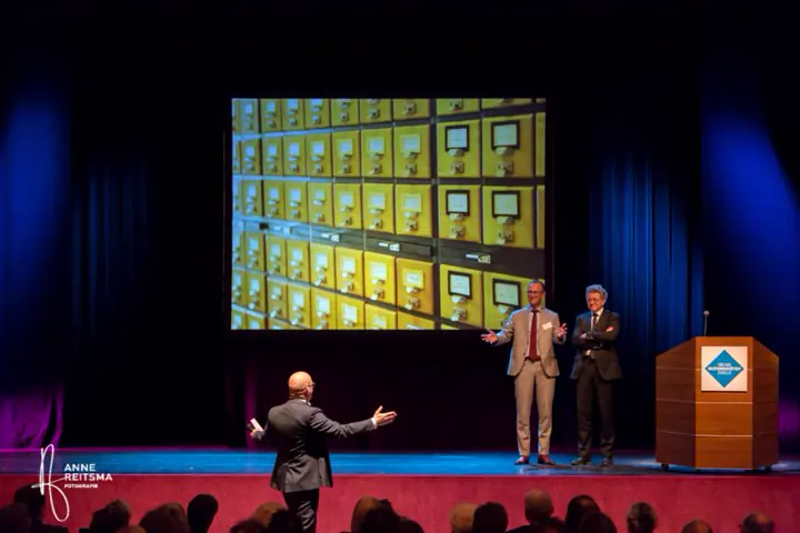 Podium met een groot scherm vol foto’s, met Gert-Jan Jansen en sprekers voor de zaal