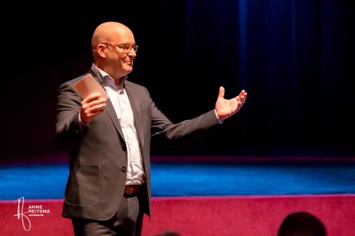 Gert-Jan Jansen spreekt met gespreide armen op een podium in blauw licht
